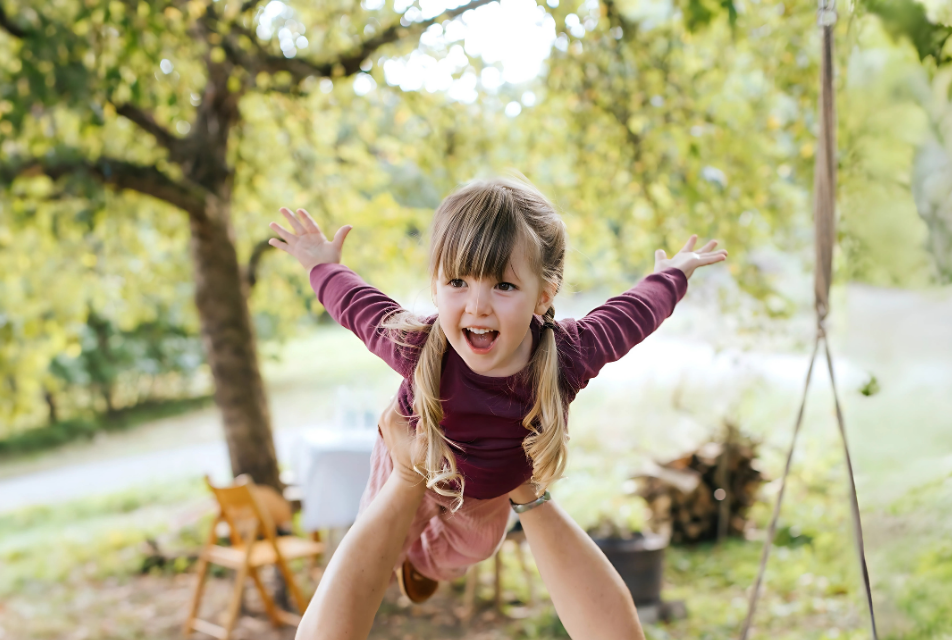 Lachendes Mädchen wird im Garten spielerisch hochgehoben – Szene voller Freude und Natur.